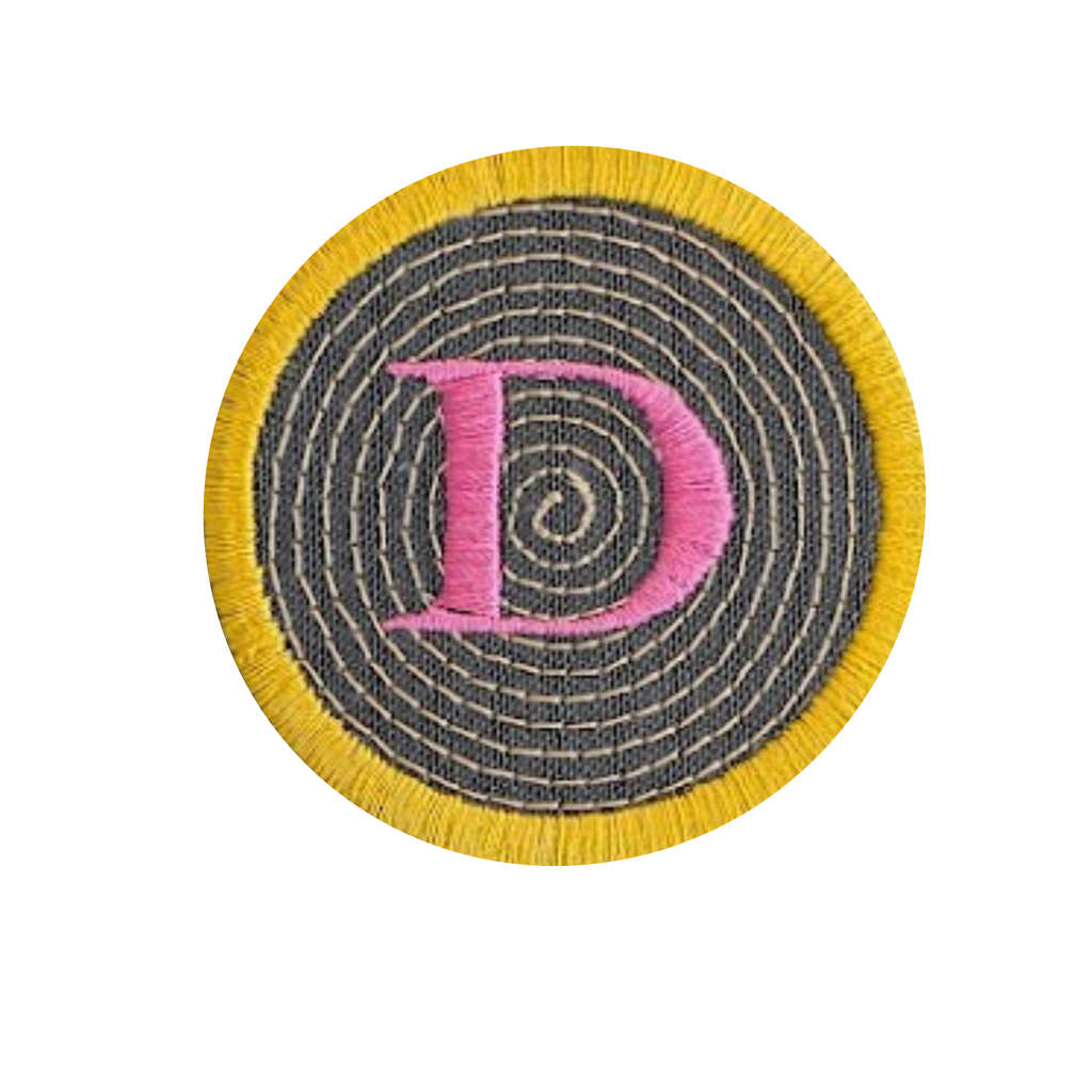 A circular embroidered patch featuring a large pink letter 'D' stitched over a dark grreen and yellow spiral pattern, surrounded by a bright yellow fringe border on a plain white background.