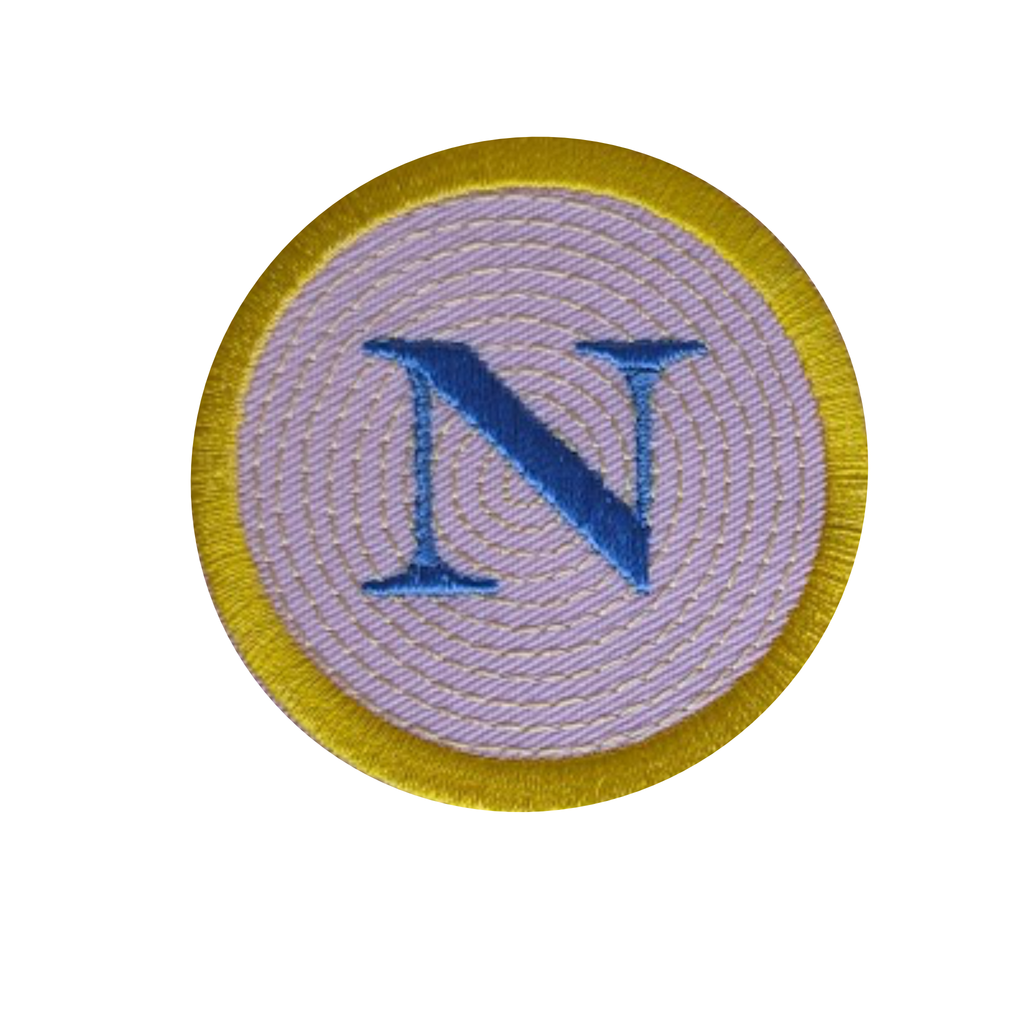 A circular embroidered patch featuring a large blue letter 'N' in the center, surrounded by a light lavender-pink stitched field and a gold border, shown on a plain white background.