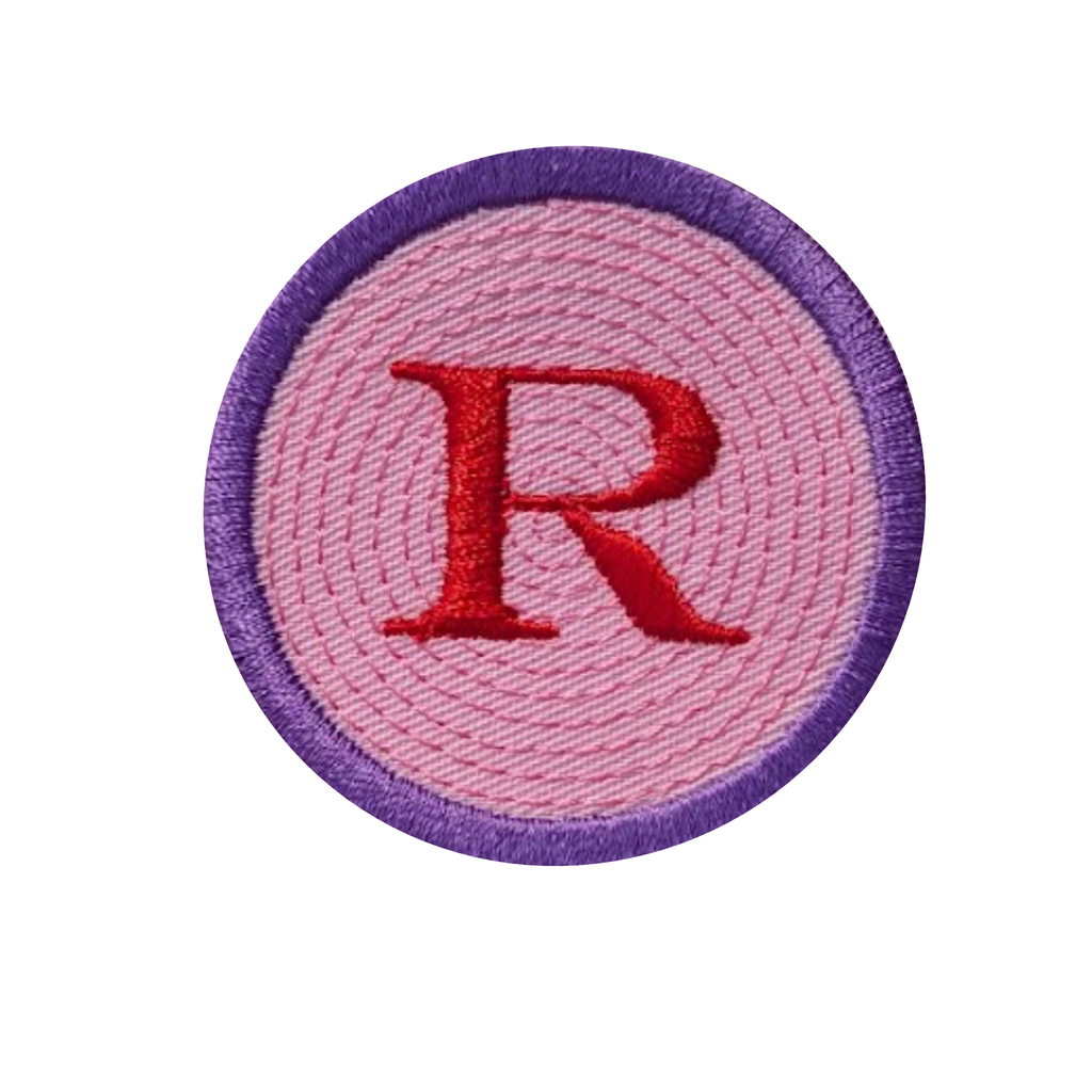 A circular embroidered patch featuring a large red letter 'R' in the center, surrounded by a pink stitched background and a purple embroidered border, shown on a plain white background.